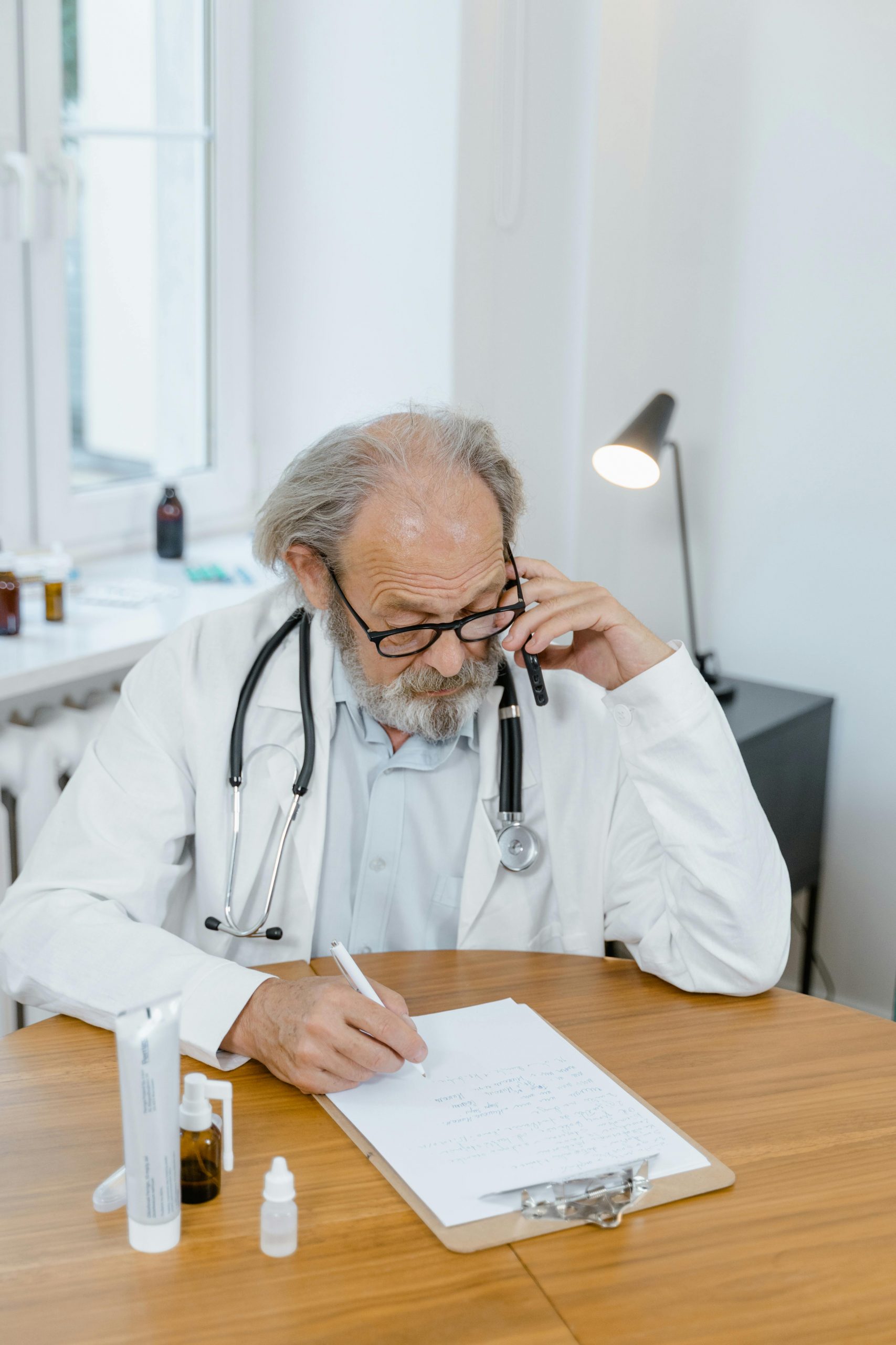 Elderly male doctor wearing glasses and a stethoscope, writing notes at a desk in a medical office.