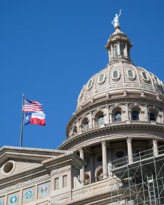 Low angle view of the Texas State Capitol dome in Austin with the American and Texas flags under a clear blue sky.